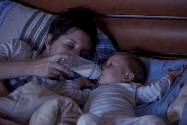 Half-asleep mom bottle feeding her infant in bed during the middle of the night