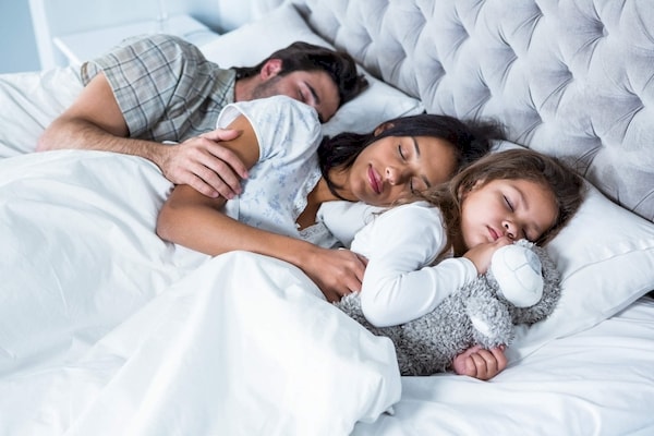 parents blissfully sleeping while sharing their bed with a toddler clutching her teddy bear